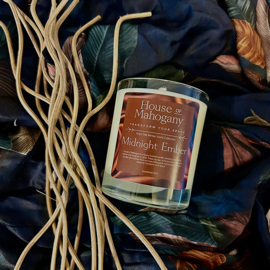 A clear candle with a label reading 'Midnight Ember' by HOUSE OF MAHOGANY LTD, surrounded by what appears to be natural soy wax with a brownish hue, on a decorative floral fabric.